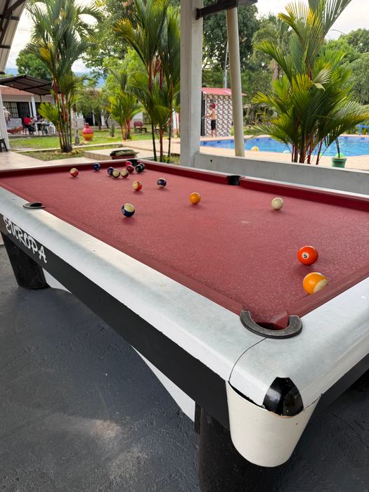 Mesa de billar de paño rojo con la piscina y las palmas al fondo