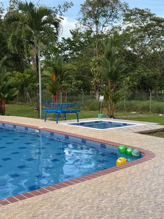 Esquina de la piscina amplia con balones y, detrás, la piscina pequeña, una banca azul y palmas